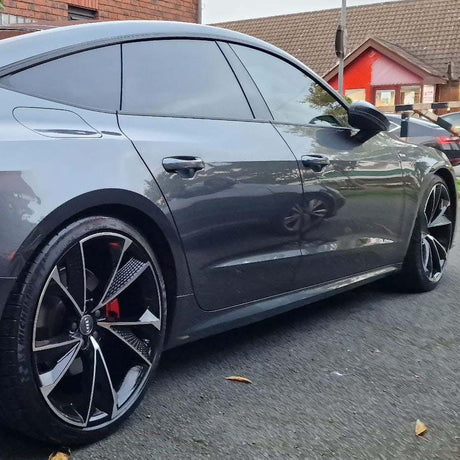 A sleek gray car is parked on the street, featuring tinted windows and stylish black alloy wheels. Nearby, there are brick and red-colored buildings, and a few scattered leaves on the ground.