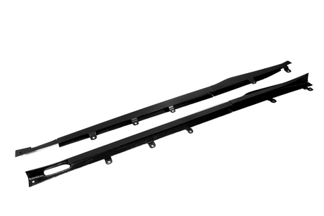 Two black carbon fiber car side skirts with attachment brackets lie against a plain white background, emphasizing their sleek design and aerodynamic purpose.