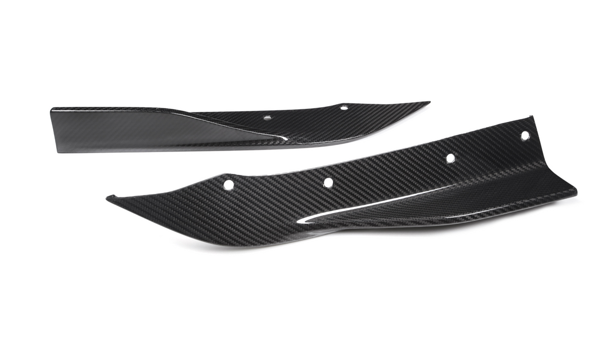 Two carbon fiber car parts, possibly splitters or fins, lie on a white background. They have aerodynamic shapes with several bolt holes for mounting.