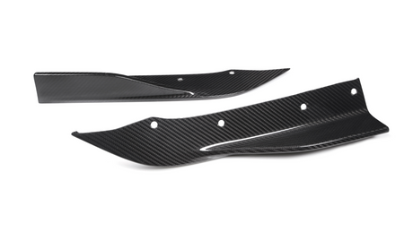Two carbon fiber car parts, possibly splitters or fins, lie on a white background. They have aerodynamic shapes with several bolt holes for mounting.