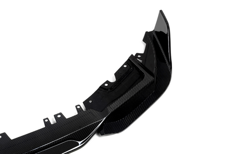 A glossy carbon fiber car accessory, likely a bumper, lies on a plain white background. The item features aerodynamic curves and multiple mounting points for attachment.