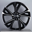 A black and silver alloy wheel features five polished spokes radiating from a central hub with the Audi logo. It's set against a plain, light gray background.