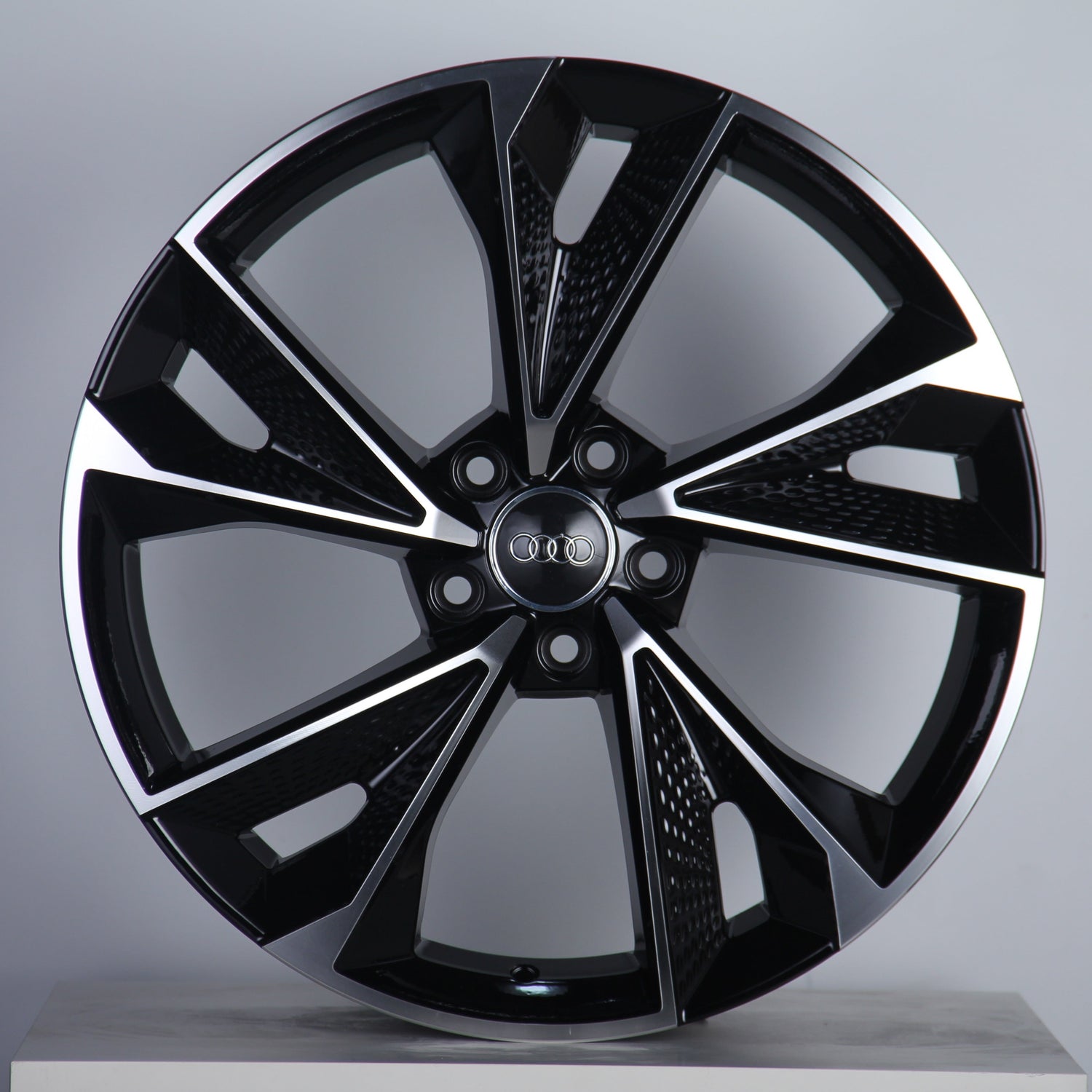 A black and silver alloy wheel features five polished spokes radiating from a central hub with the Audi logo. It's set against a plain, light gray background.