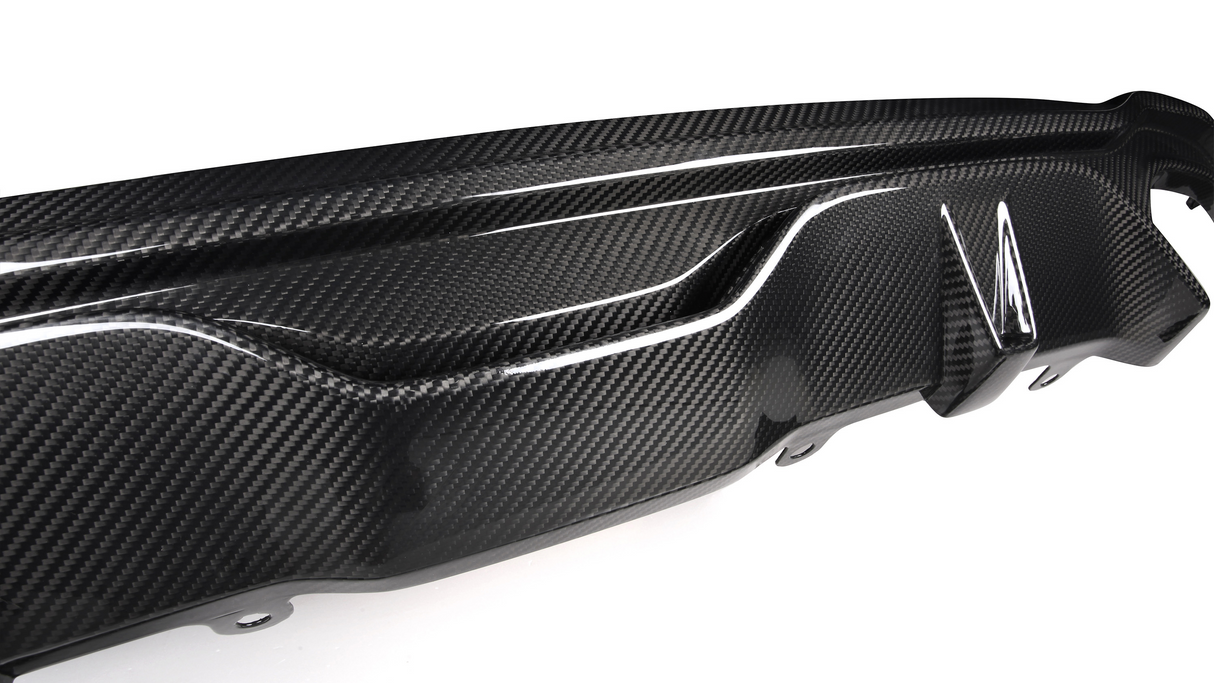 A carbon fiber car bumper diffuser features a textured design, showcasing glossy, aerodynamic curves and fins, mounted on a white surface, emphasizing its sleek automotive styling.