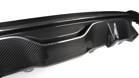 A carbon fiber car bumper diffuser features a textured design, showcasing glossy, aerodynamic curves and fins, mounted on a white surface, emphasizing its sleek automotive styling.