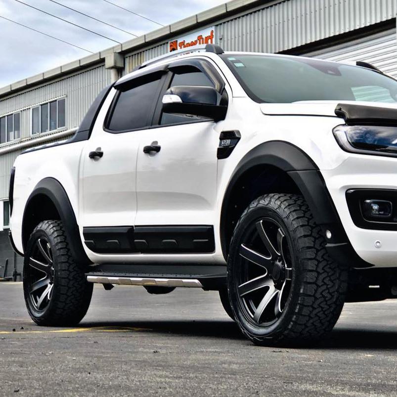 A white pickup truck with large black wheels is parked in front of a corrugated metal building under overcast skies. A sign on the building reads "FleetFirst."