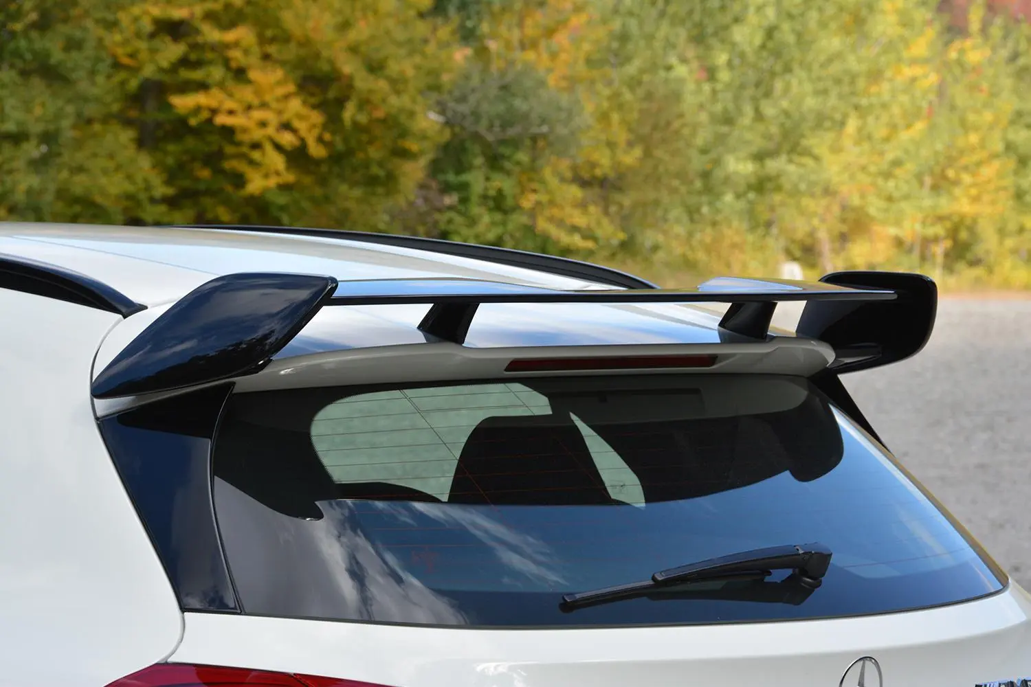 A sleek car spoiler perched on the rear of a white vehicle, situated against a backdrop of green and yellow foliage on a clear day.