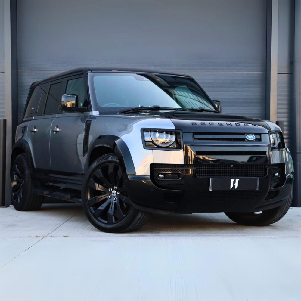 A grey Land Rover Defender with large black wheels is parked on a concrete surface in front of a closed grey garage door. "DEFENDER" is visible on the vehicle’s front.