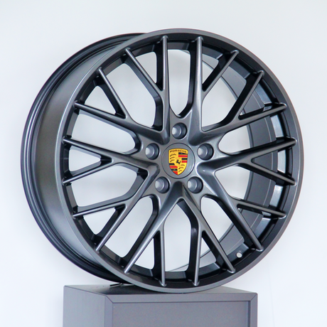 A dark alloy wheel with a multi-spoke design and a central Porsche logo is displayed upright against a plain white background.