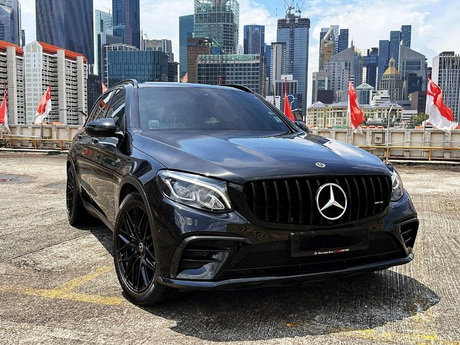 A black Mercedes-Benz SUV is parked on a rooftop, surrounded by Singapore city skyscrapers and red flags. The grill features the Mercedes logo prominently.