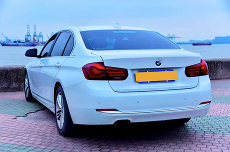 A white car is parked on a paved area near a waterfront. Cargo ships and cranes are visible in the background across the water.