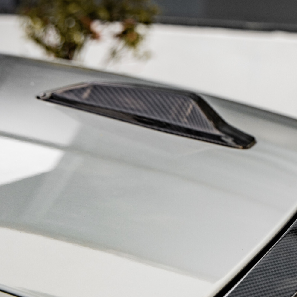 A carbon fiber vent is mounted on a smooth, light-colored car hood, situated in an outdoor setting with blurred foliage visible in the background.