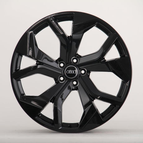 A shiny black, multi-spoke alloy wheel with an "Audi" emblem in the center is displayed against a plain, light gray background.