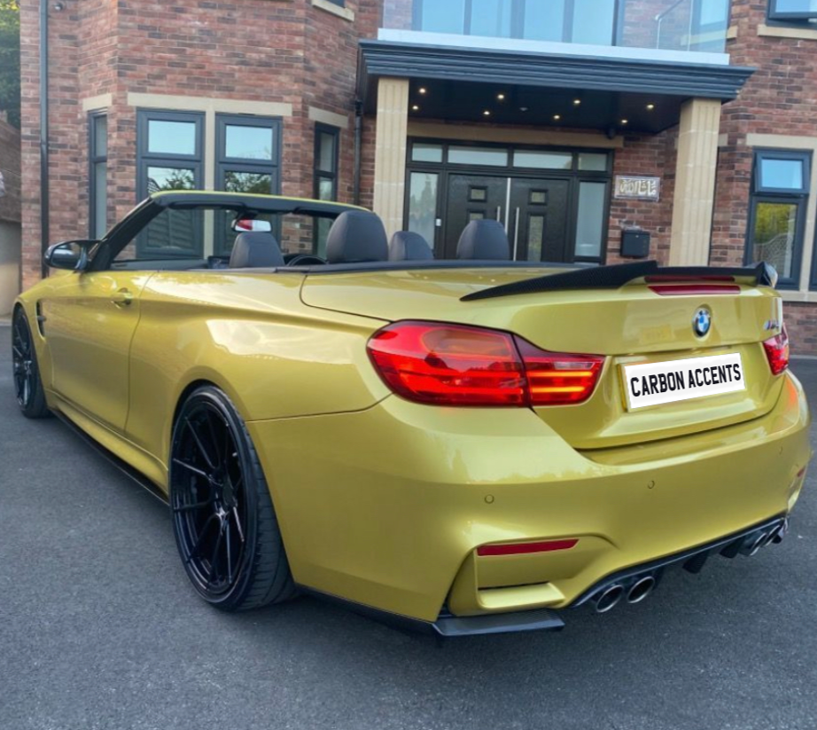 Convertible gold car with black wheels parked in front of a brick house with large windows and a double-door entrance. License plate reads "CARBON ACCENTS."
