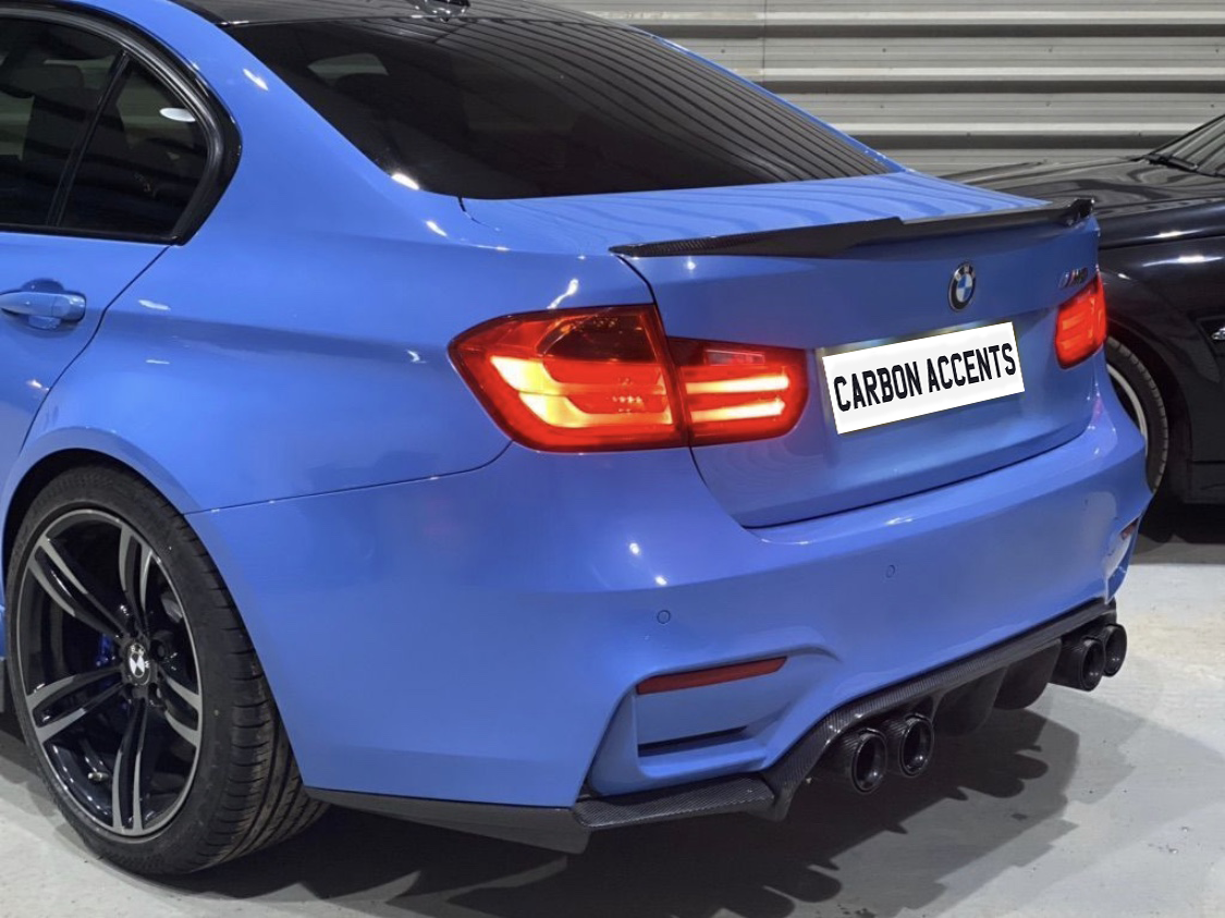 A blue car with illuminated taillights is parked in an indoor garage. The license plate reads, "CARBON ACCENTS." The car has dual exhausts and a carbon-fiber rear spoiler.