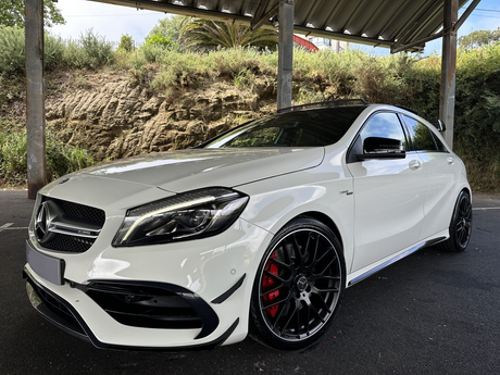 A white Mercedes-Benz sedan is parked under a metal-roofed structure, featuring sleek black wheels and set against a backdrop of rocks and dense shrubs.