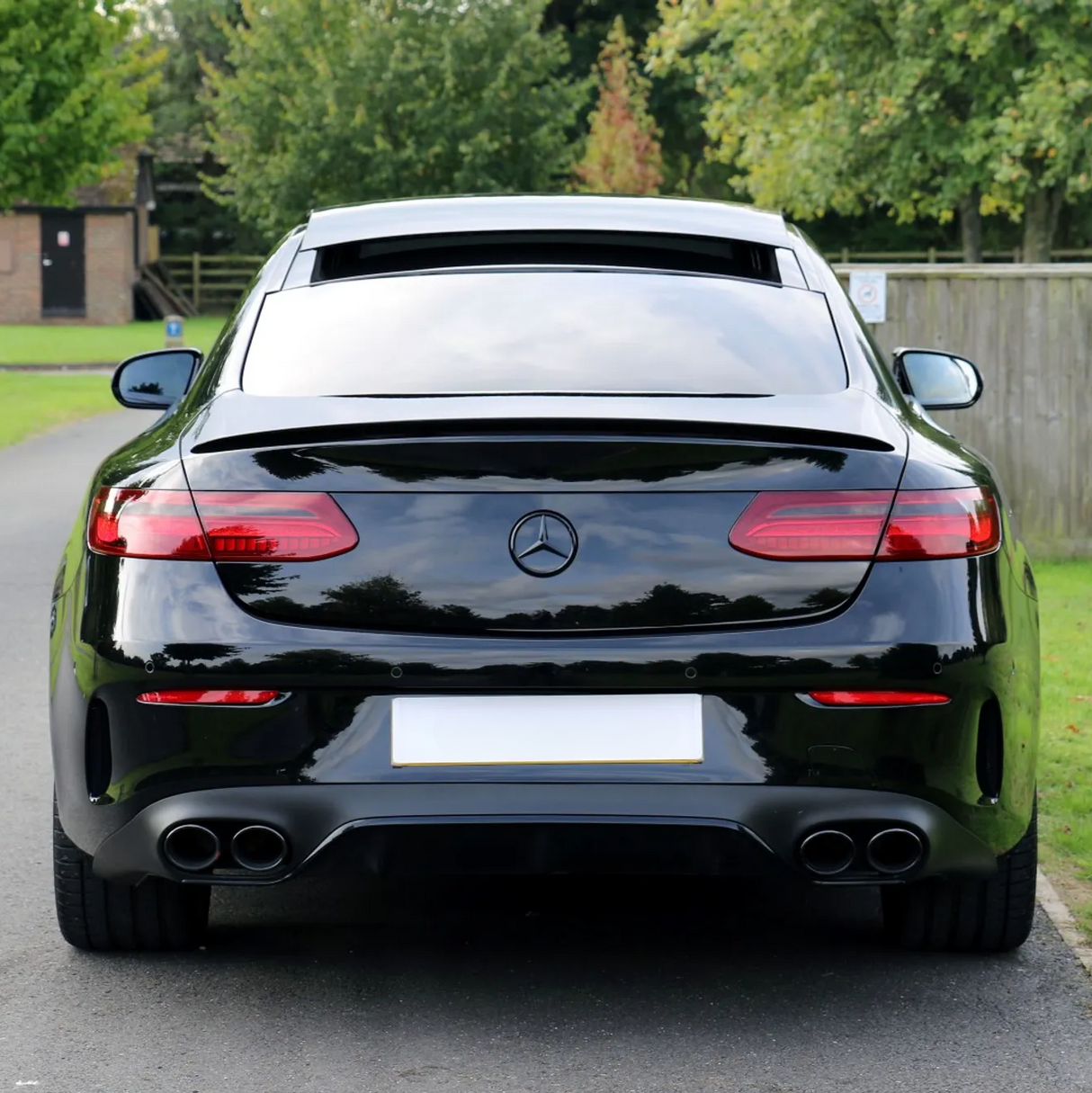 A sleek black Mercedes-Benz car is parked on a paved road, surrounded by lush greenery and wooden fencing in a suburban area.