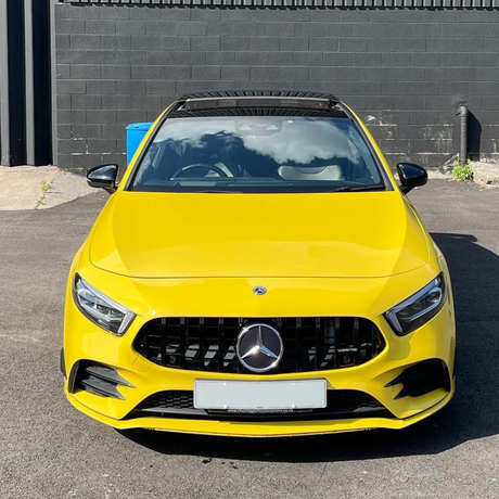 A bright yellow Mercedes-Benz parked on an asphalt surface, reflecting clouds on its windshield, is positioned against a black brick wall. A blue bin is partially visible beside it.