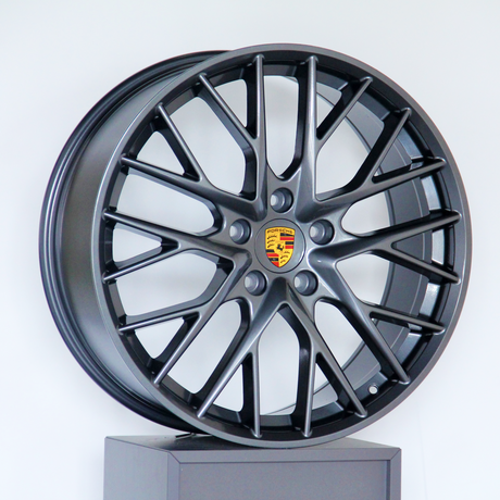 A dark metallic alloy wheel with multiple spokes and a central emblem featuring the Porsche logo, displayed on a platform against a plain white background.