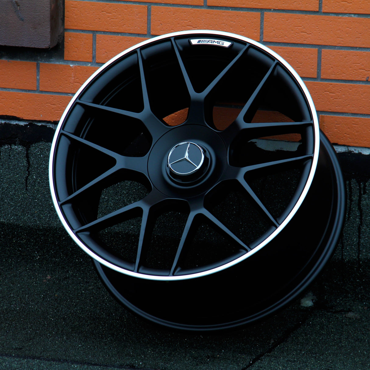 A black Mercedes-Benz AMG wheel rests against a brick wall on a dark, textured ground. The wheel features a silver trim and the Mercedes-Benz logo at the center.