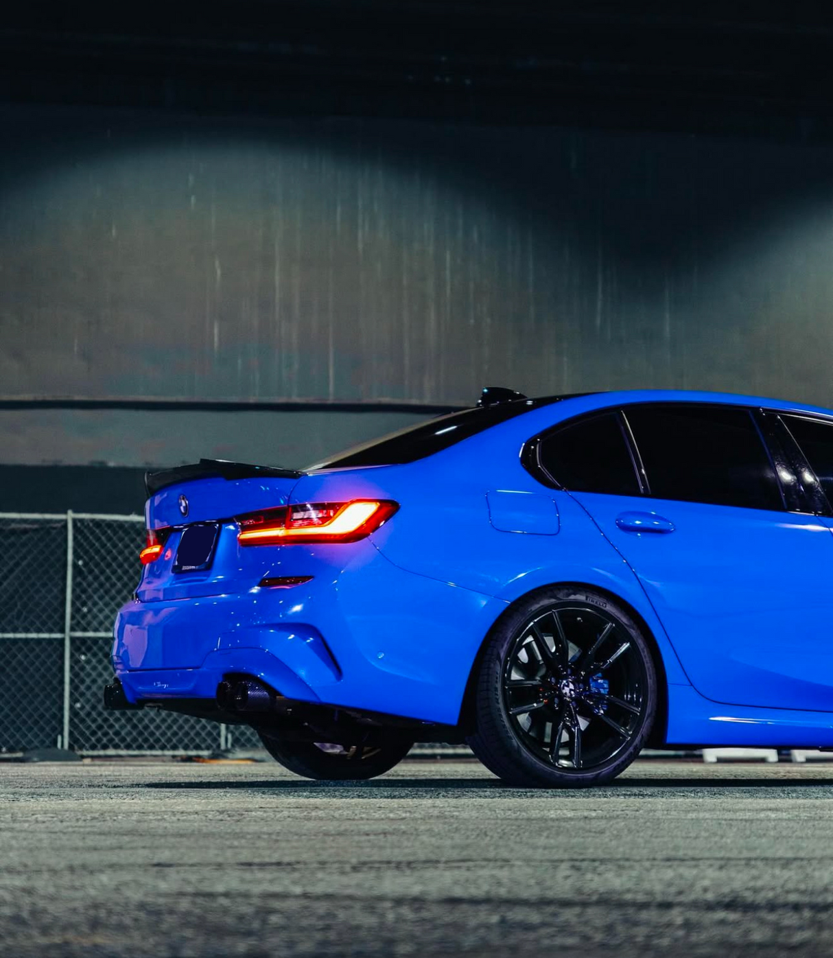 A blue sedan with illuminated taillights is parked on a concrete surface, surrounded by a chain-link fence and under an industrial-style structure.
