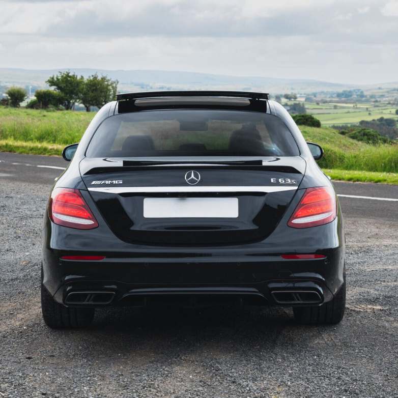 A black Mercedes-Benz E63S is parked on a paved road, with green fields and a cloudy sky in the background. Visible text: "AMG", "E 63 S".