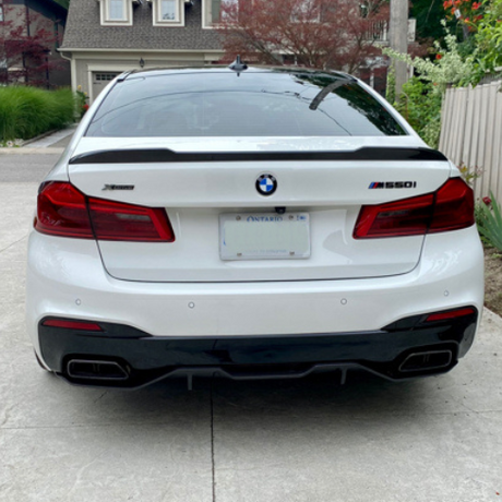 A white BMW 550i is parked in a driveway, displaying a license plate from Ontario. The car features a black rear diffuser and is set against a residential background with greenery.