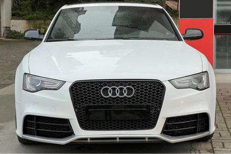Car: A white Audi with rain droplets is parked in front of a red building or structure, surrounded by greenery and paving stones.