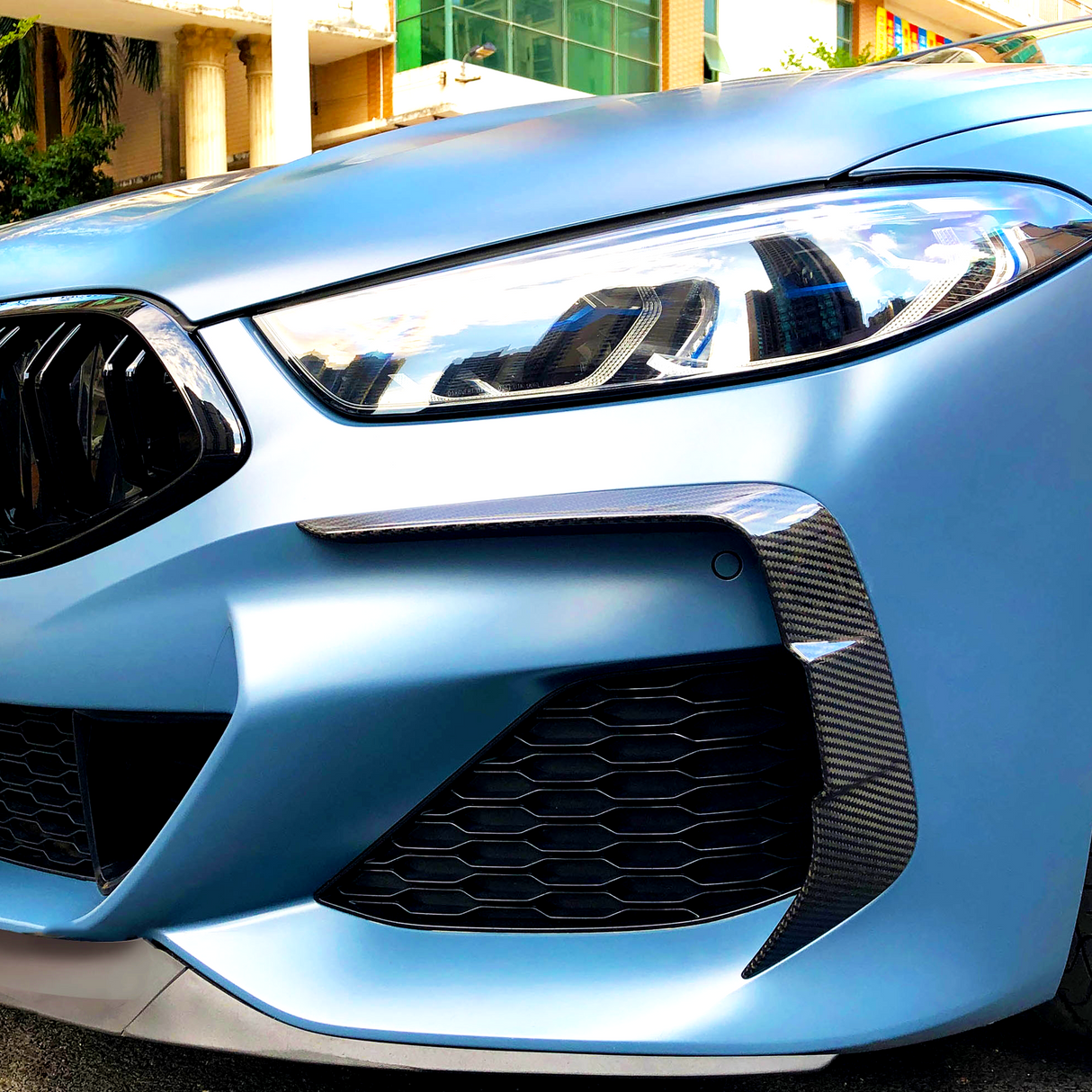A sleek car front features angular headlights and a sporty grille, parked in an urban environment with modern buildings in the background.