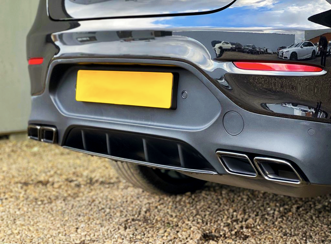 The rear end of a dark car is parked on gravel, featuring a bright yellow license plate, dual exhausts, and red reflectors. Nearby, other vehicles are visible in the reflection.