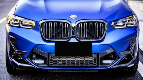 A blue car's front features a modified grille with a smiling logo, surrounded by bright headlights. The vehicle is parked against a blurred urban background.