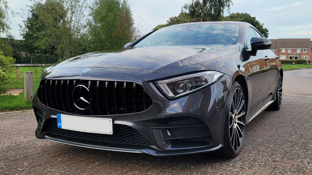 A sleek dark gray Mercedes-Benz car is parked on a brick driveway. The surrounding area features green grass, trees, and a few houses in the background.