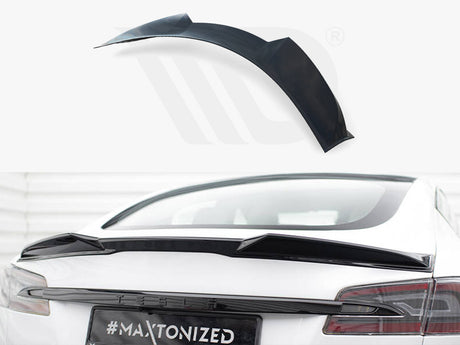 A black car spoiler is positioned on the rear of a white vehicle, with the text "#MAXTONIZED" on the license plate. It rests in a bright, indoor environment.