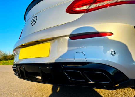 A silver car's rear bumper with a yellow license plate and dual exhaust is parked on a road. The context includes a clear blue sky and sunlight casting shadows.