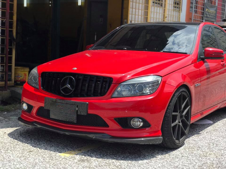 Red Mercedes-Benz car parked on a sunlit street, featuring black rims and tinted windows. Nearby, there are buildings with yellow grids and a partially open garage door.