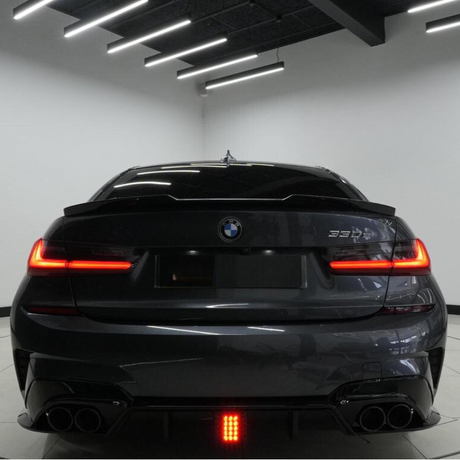 A dark-colored BMW 330i is parked indoors, showing its illuminated rear lights. The sleek, modern ceiling features hanging fluorescent lights, adding a contemporary ambiance to the garage setting.