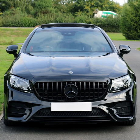 Car stationary on a road; glossy black body, wide grille with a logo, surrounded by lush green trees and grass. License plate is blank.