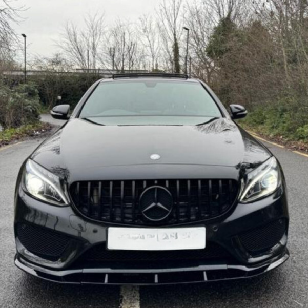A black Mercedes car is parked on a road lined with leafless trees and bushes. It has illuminated headlights and a visible Mercedes emblem at the front center.