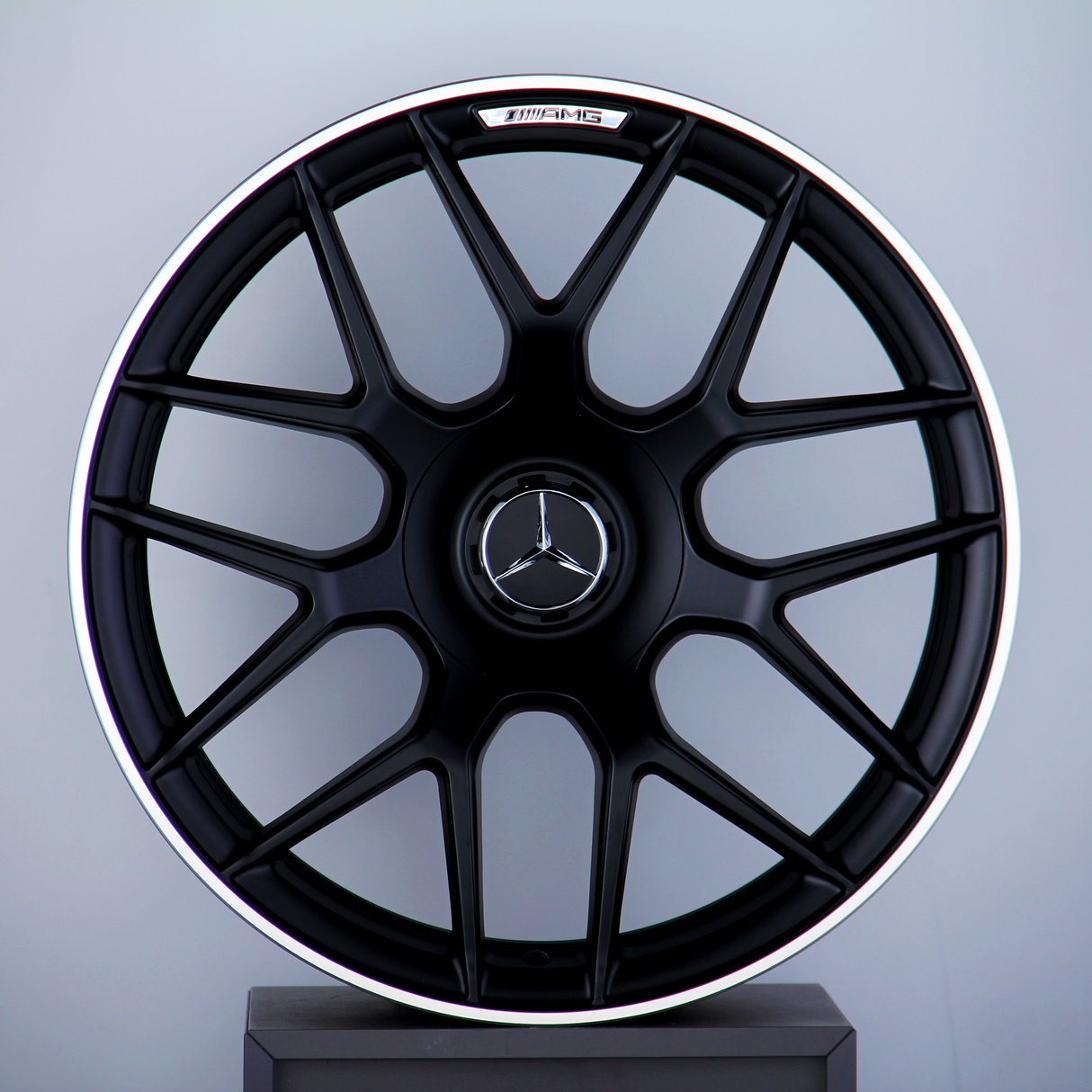 A black Mercedes-AMG wheel with a multi-spoke design stands upright against a gray background. The Mercedes logo is centered, with "AMG" text at the top of the wheel.