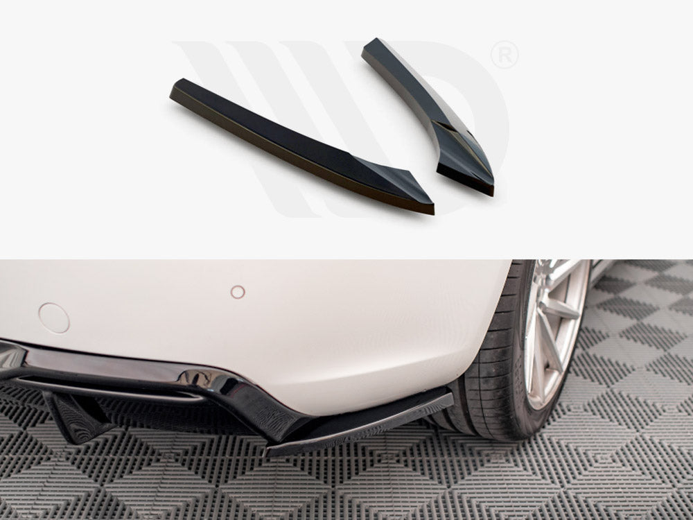 Two black car diffuser fins rest on a white surface above a close-up of a car's rear bumper with a diffuser installed, parked on a patterned grey floor.