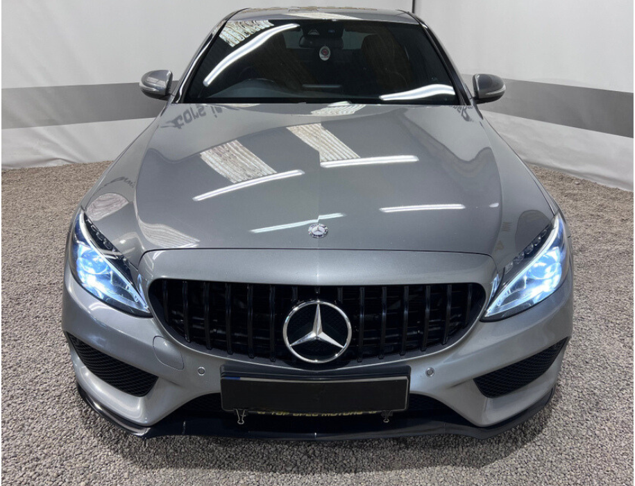 A silver car with illuminated headlights is parked on a gravel floor, positioned against a light-colored backdrop.