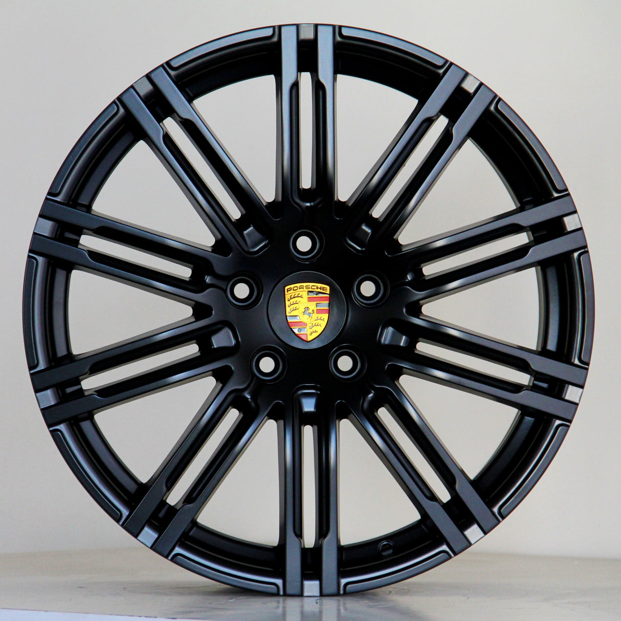A black alloy wheel with multiple spokes features a central emblem displaying the Porsche logo. The wheel is positioned against a plain light gray background.