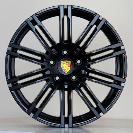 A black alloy wheel with multiple spokes features a central emblem displaying the Porsche logo. The wheel is positioned against a plain light gray background.