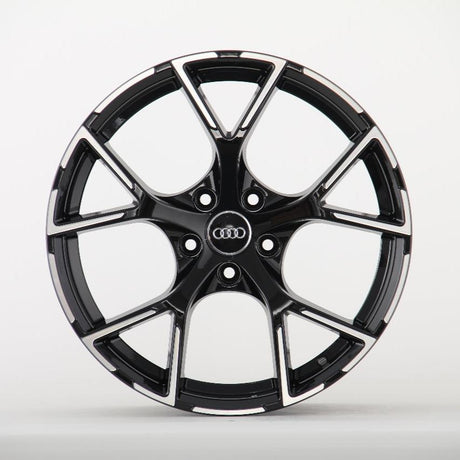 A black alloy wheel with a shiny finish features the Audi logo at its center, set against a plain white background. The wheel has multiple spokes and a sporty design.