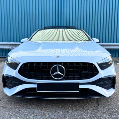 A silver car with a prominent front grille and logo is parked, facing forward on a gravel surface, set against a corrugated blue metal wall backdrop.