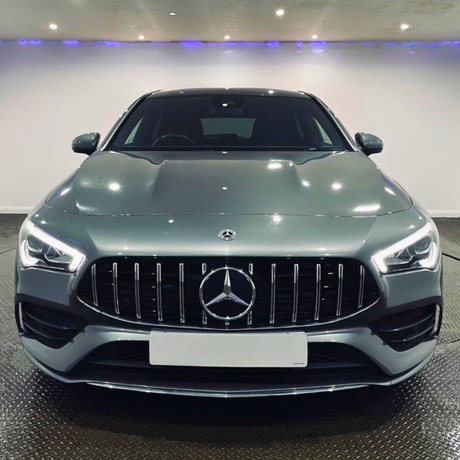 A silver car with prominent grille and illuminated headlights is parked in a brightly lit showroom with a white wall and patterned floor.