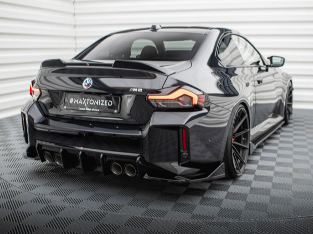 A black BMW M2 with large rims and quad exhaust parked on a checkered floor in a white-paneled room. License plate: "#MAXTONIZED" and below, "Dealer Maxton de."