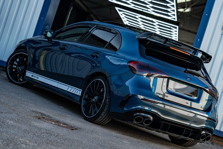 A blue Mercedes-Benz AMG car is parked, showcasing its sleek design and rear spoiler. It's situated in front of a building with illuminated patterns above.