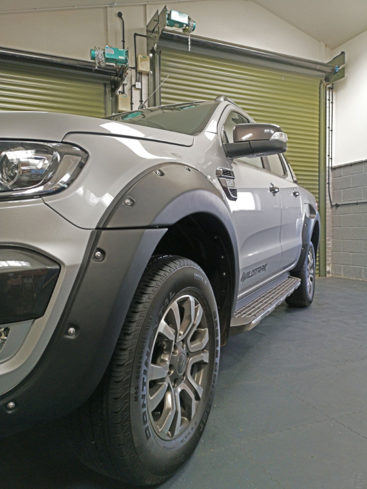 A silver pickup truck parked inside a garage with green roll-up doors visible. "Wildtrak" is written on the side. The image highlights the front wheel and fender.