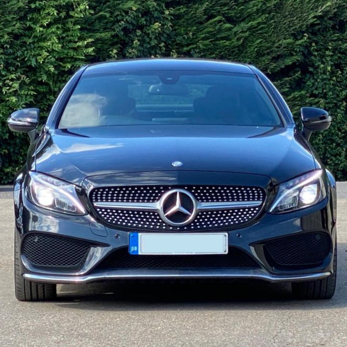 A black Mercedes-Benz car parked on a paved surface, facing forward, surrounded by green foliage. The front grille features the iconic emblem, with the license plate below.